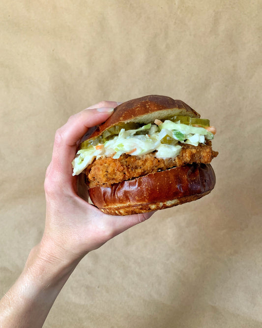 Hand holding up a Crispy Seitan Cutlet burger with slaw and pickles